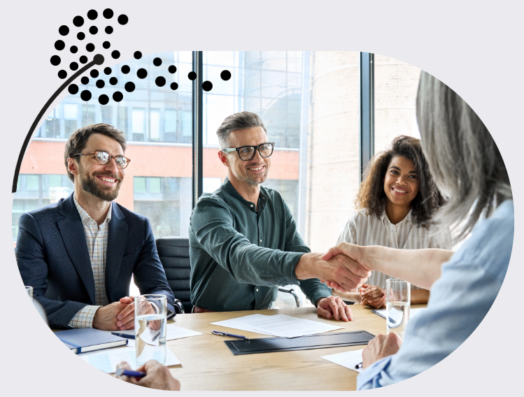 The image shows four people sitting in a meeting. There are two white men and a black woman forward facing. The man in the middle is shaking the hand of a white women with grey hair sitting opposite to him. The image is set against a light grey background and a black dandelion decorate the top-left corner of the image.